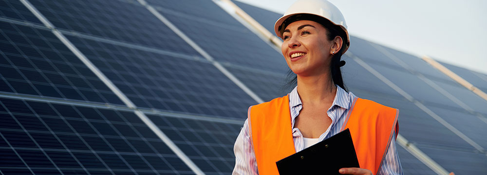 Mujer con casco blanco y chaleco naranja sonriendo, frente a paneles solares